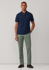 Polo bleu marine avec bordure blanche, deux boutons au col, associé à un pantalon vert clair et des chaussures marron foncé. Design simple, texture lisse.