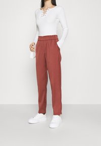 Witte lange mouwen top met een vetersluiting bij de hals, gecombineerd met losse roestkleurige broek met een elastische tailleband en zijzakken. Witte sneakers.