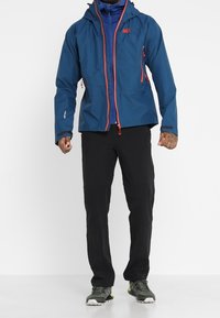 Giacca blu impermeabile con dettagli rossi, cappuccio regolabile, due tasche con zip; abbinata a pantaloni da esterno neri e scarpe sportive grigio-verdi.