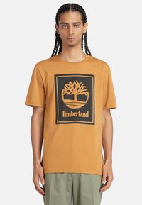 Košile s krátkým rukávem, oranžová bavlněná, s černým logem stromu a textem "Timberland" v obdélníkovém designu na přední straně.