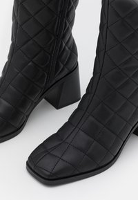 Bottines noires matelassées à bout carré, en cuir synthétique texturé, avec fermeture éclair sur le côté et un talon empilé modéré.