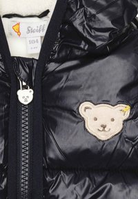 Giubbotto imbottito blu navy con finitura lucida, dotato di una patch con la testa di un orso sul lato sinistro e di un tirazip a forma di orso. Fodera interna morbida.