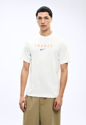 Fiatal férfi fehér "FRANCE" feliratú és kék Nike logós pólóban, bézs nadrággal, egyszínű háttér előtt állva.