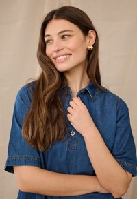 Fiatal nő hosszú barna hajjal, arany fülbevalóval és kék farmeringben, mosolyogva oldalt nézve, karjait keresztbe téve.