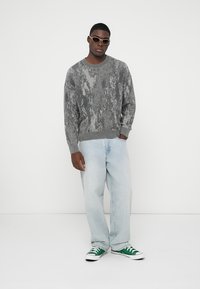 Γκρίζα, υφαντή πλεκτή μπλούζα με σχέδιο, συνδυασμένη με ανοιχτό μπλε φαρδιά τζιν και πράσινα και λευκά αθλητικά παπούτσια.