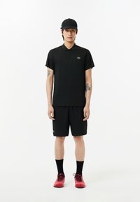 Svart polo tröja med korta ärmar och grön logotyp, matchad med svarta shorts och röda träningsskor. Tillbehör inkluderar en svart keps.