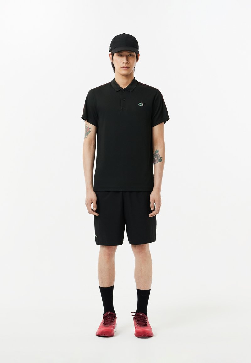 Schwarzes Polohemd mit kurzen Ärmeln und grünem Logo, kombiniert mit schwarzen Shorts und roten Sportschuhen. Zubehör umfasst eine schwarze Kappe.