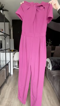 Fuchsia jumpsuit met korte mouwen en een strik aan de hals. Heeft een brede broekspijp en plooien, gemaakt van een glad, lichtgewicht materiaal.
