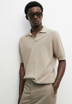 Uomo con polo in maglia beige a maniche corte e pantaloni abbinati, con occhiali da sole rettangolari scuri, in piedi su sfondo bianco.