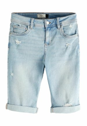 Femme portant un chemisier en dentelle blanche et un short en jean bleu clair déchiré, debout sur une plage rocheuse avec l'océan en arrière-plan.