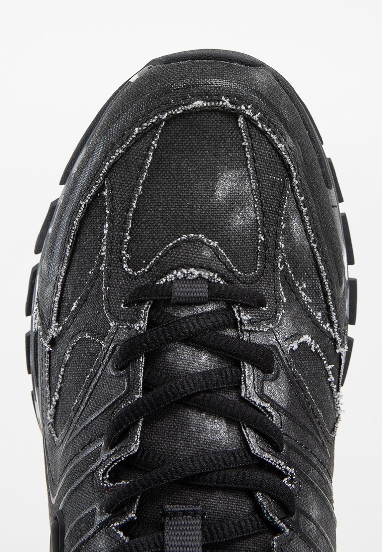 Chaussure de sport noire avec une tige en tissu texturé, des coutures contrastantes et une semelle rembourrée. Dispose d'un design à lacets et d'accents en caoutchouc.