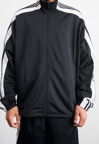 Svart zip-up jacka i ett lätt och glatt material med vita sidostripes, hög krage och elastiska mudd.