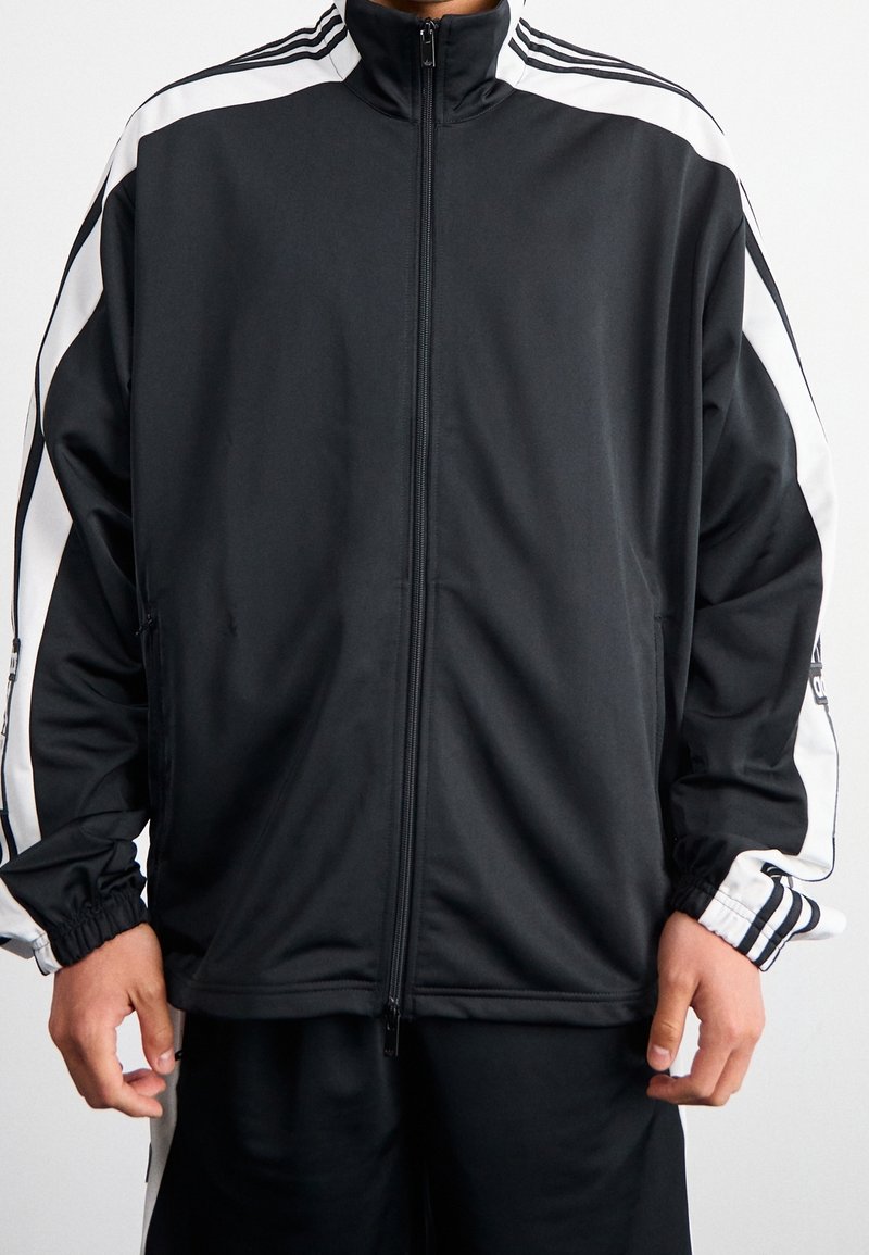 Svart zip-up jacka i ett lätt och glatt material med vita sidostripes, hög krage och elastiska mudd.