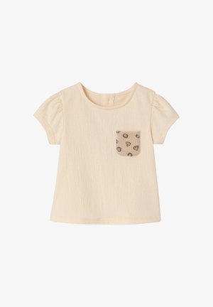 Babyschirt in Creme mit kurzen Ärmeln, strukturierter Stoff und eine kleine beige Tasche mit dunklen Herz- und Wirbelmustern auf der Vorderseite.