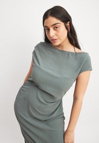 Draped, short-sleeve dress in muted green fabric with a soft texture and asymmetrical neckline, featuring a fitted silhouette.