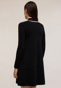 Abito nero a maniche lunghe con una vestibilità comoda, dotato di un colletto alto e dettagli crema sulle spalle. Tessuto liscio, lunghezza al ginocchio.