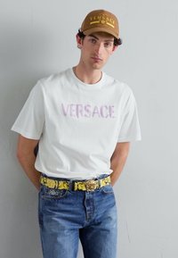 Weißes Kurzarm-T-Shirt mit pastellfarbenem "VERSACE"-Logo, blaue Jeans und einem gelben Blumen-Gürtel mit goldener Schnalle. Braune Mütze mit Logo.