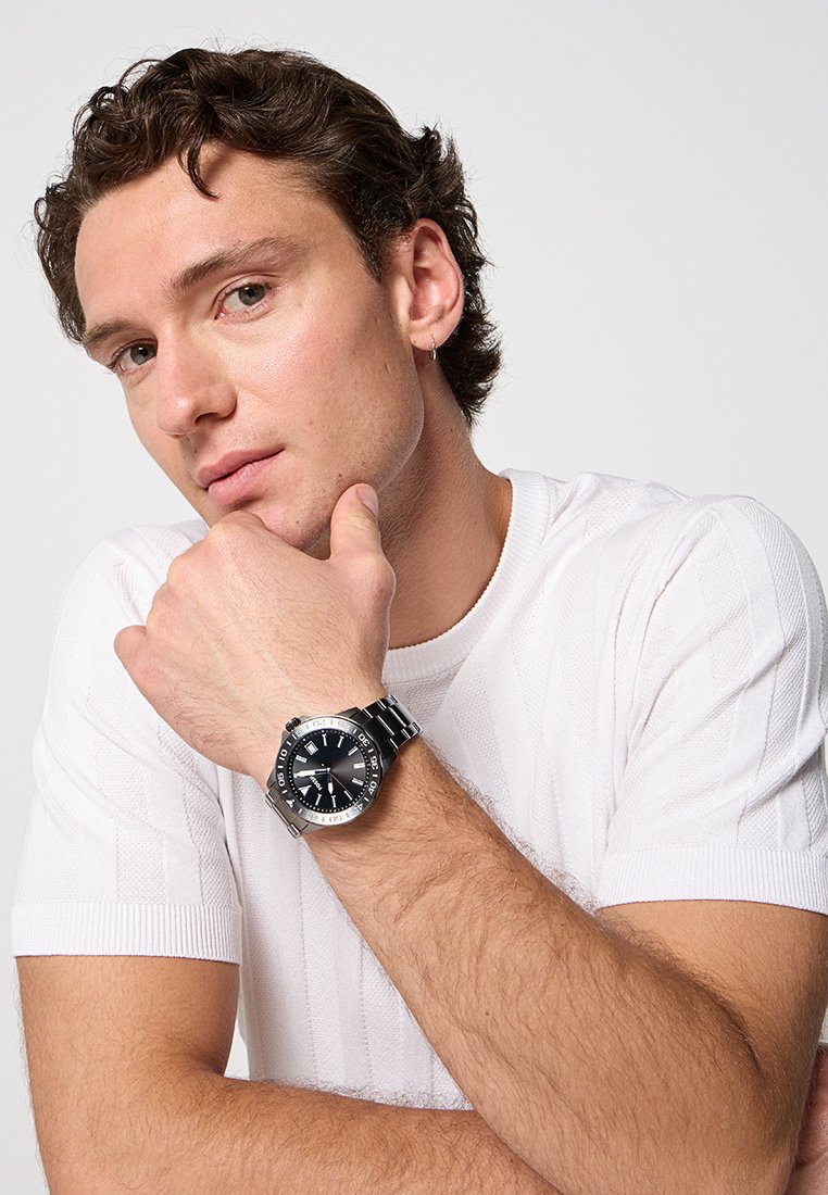 Orologio in acciaio inox argento con quadrante nero, indicatori luminosi e lunetta girevole indossato a un polso, con una camicia bianca a trama visibile.