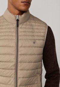 Chaleco acolchado beige con cuello alto, mangas marrones en contraste, cierre de cremallera delantero y un pequeño logo negro en el pecho.
