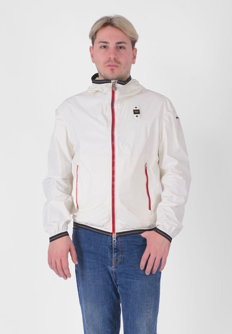 Giacca leggera bianca con zip frontale e accenti rossi, dotata di colletto alto e polsini elastici. Indossata sopra jeans blu.