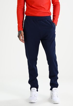 Mężczyzna w granatowych spodniach dresowych, czerwonej bluzie z długim rękawem i białych trampkach, stojący z jedną ręką w kieszeni na białym tle.