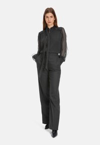 Schwarze Bluse aus durchsichtiger Stoff mit Polka-Dots und Bindegürtel, lange Puffärmel; hochtaillierte schwarze Hose mit taillierter Passform; spitze schwarze Ankle Boots.