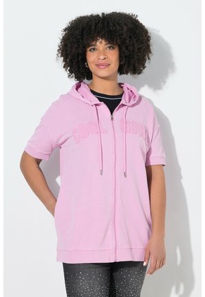 Kvinde med krøllet hår iført en lyserød kortærmet hættetrøje med teksten "COOL GIRL" og sorte bukser med pletter, der står foran en hvid baggrund.