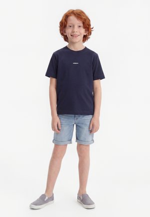 Dreng med rødt hår iført en navy t-shirt, lyseblå op rullede denimshorts og grå slip-on sko, står og smiler.