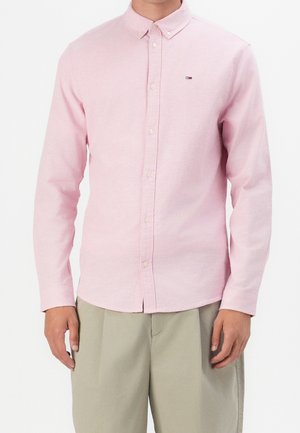 Homme portant une chemise rose clair à manches longues boutonnée et un pantalon beige, debout devant un fond blanc uni.