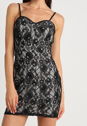 Femme portant une mini robe noire en dentelle sans manches avec des motifs floraux et des fines bretelles sur un fond uni.