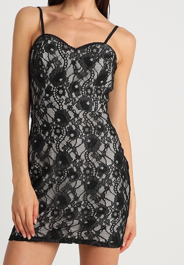 Femme portant une mini robe noire en dentelle sans manches avec des motifs floraux et des fines bretelles sur un fond uni.