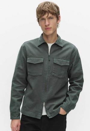 Ung man med ljust brunt hår som bär en mörkgrön jacka med dragkedja och två bröstfickor, stående mot en enkel vit bakgrund.