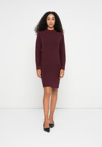 Donna con capelli ricci che indossa un vestito in maglia borgogna a maniche lunghe e lunghezza al ginocchio, con tacchi neri a punta, in piedi su un pavimento bianco.