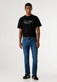 Mladý muž stojící v černém tričku Pepe Jeans, modrých džínách a černých teniskách na jednoduchém světlém pozadí.