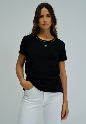 Mujer con camiseta negra de manga corta con pequeño logo blanco en el pecho, pantalones blancos, de pie con una mano en el bolsillo contra un fondo liso.