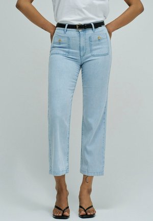 Vaqueros cortos de tiro alto en azul claro con bolsillos delanteros de parche y detalles de botones, combinados con un cinturón negro y tacones abiertos negros.