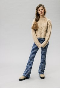 Beige gebreide trui met een geweven ruitpatroon en een losse pasvorm, gecombineerd met blauwe gestreepte broek met een druppelontwerp en zwarte laarzen.