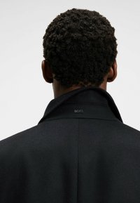Manteau en laine noir avec un col haut, présentant une étiquette « BOSS » discrète au dos. La texture semble lisse et sur mesure.