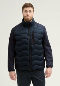 Veste matelassée navy avec design en piquage, col haut, manches en tissu contrasté et poches zippées. Présente une texture lisse et mate.