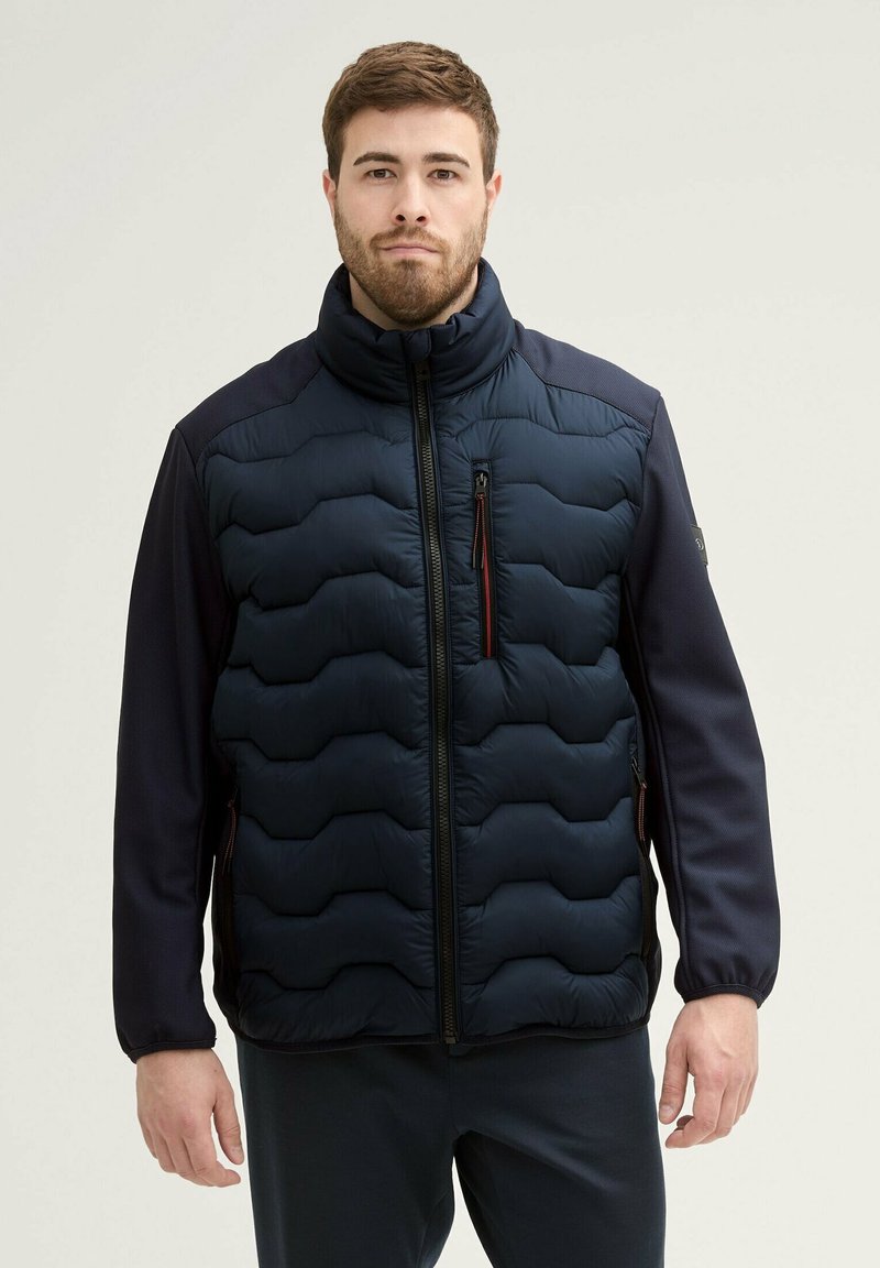 Veste matelassée navy avec design en piquage, col haut, manches en tissu contrasté et poches zippées. Présente une texture lisse et mate.