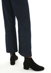 Botas de tobillo de ante negro con un tacón bajo y cuadrado, con una cremallera lateral, combinadas con pantalones de mezclilla oscuros de pierna ancha con dobladillo sin rematar.