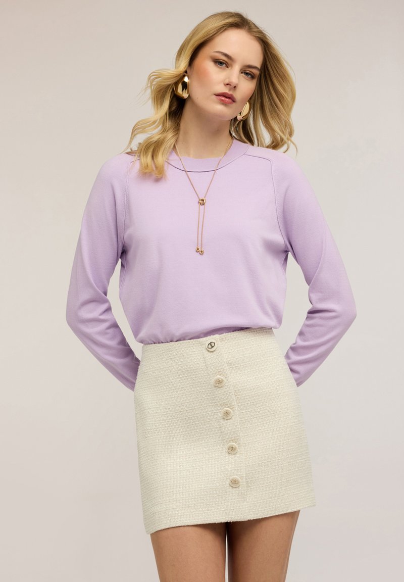 Femme portant un haut à manches longues lavande rentré dans une mini-jupe blanche texturée à boutons sur le devant, accessoirisée avec des boucles d'oreilles dorées et un collier pendentif.