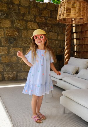 Gestreiftes blau-weißes Kleid mit bunter Stickerei, gerafftem Saum, Strohhut, pinken runden Sonnenbrillen und pinken Sandalen. Hintergrund aus Steinmauer.