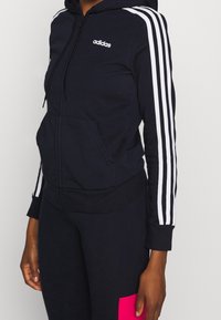 Sudadera con capucha de algodón en mezcla azul marino con cremallera; presenta detalles de tres rayas blancas en las mangas y el logo frontal. Bolsillo tipo canguro en la parte delantera.