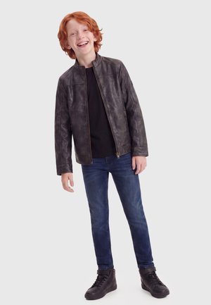 Ragazzo dai capelli rossi che sorride, indossa una giacca di pelle nera, una maglietta nera, jeans blu e sneakers alte nere, in piedi su uno sfondo bianco.