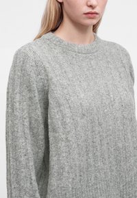 Jeune personne aux longs cheveux blonds portant un pull en laine gris clair côtelé, visible des épaules au menton sur un fond uni.