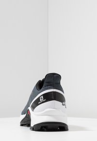 Donkerblauwe hardloopschoen met een gestructureerde bovenkant, zwarte accenten, een witte tussenzool en een robuuste outsole. Heeft merkvermelding aan de zijkant en hiel.