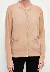 Cardigan en tricot beige avec un col rond, fermeture à bouton et deux poches avant. Présente des poignets et un ourlet côtelés. Texture lisse.