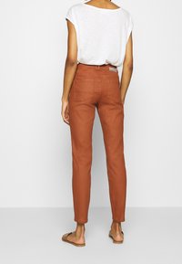 Rust-colored cotton pants with a slim fit, back pockets, and a mid-rise waist, paired with a loose white top and flat sandals.