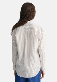 Biała koszula zapinana na guziki z granatowymi pionowymi paskami, z kołnierzykiem typu spread, długimi rękawami i zaokrąglonym dołem. Wykonana z miękkiej bawełny.